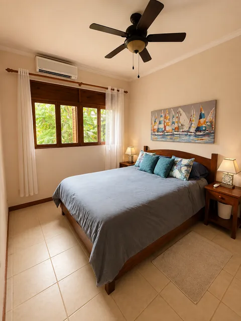 Bedroom 2 with a queen bed in teal and blue upholstery, sailboat artwork, and a jungle-view window with air conditioning